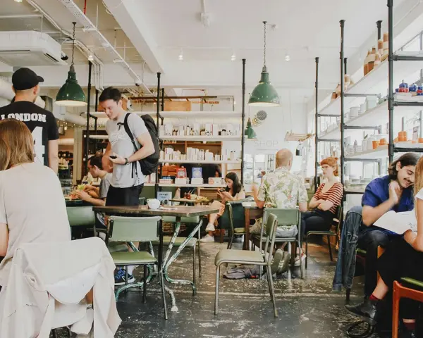 Gente en un café con mesas verdes y estanterías llenas de objetos. El **ruido restaurante** es palpable con las conversaciones y el movimiento.