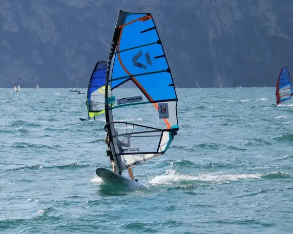 Osoba na desce windsurfingowej z niebieskim żaglem na wzburzonym morzu.