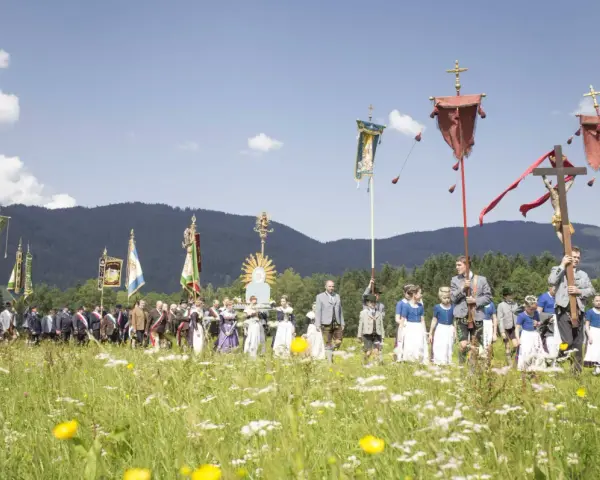 Menschen in Tracht und Uniformen nehmen an einer Prozession teil, die den Fronleichnam feiert. Fahnen und ein Kreuz werden getragen.