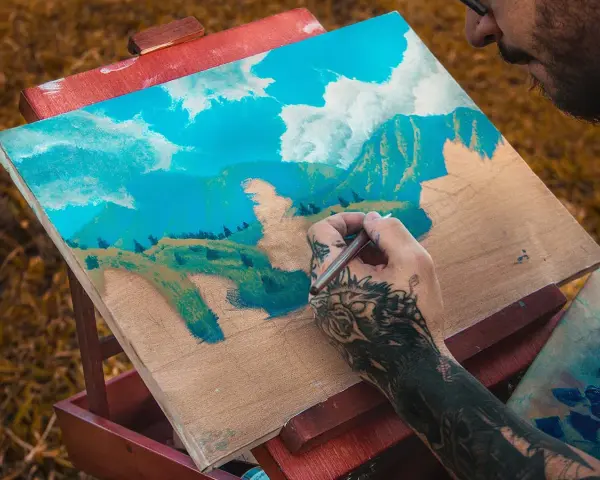 Un artiste tatoué est en train de peindre à la gouache sur toile un paysage montagneux sous un ciel bleu et nuageux.