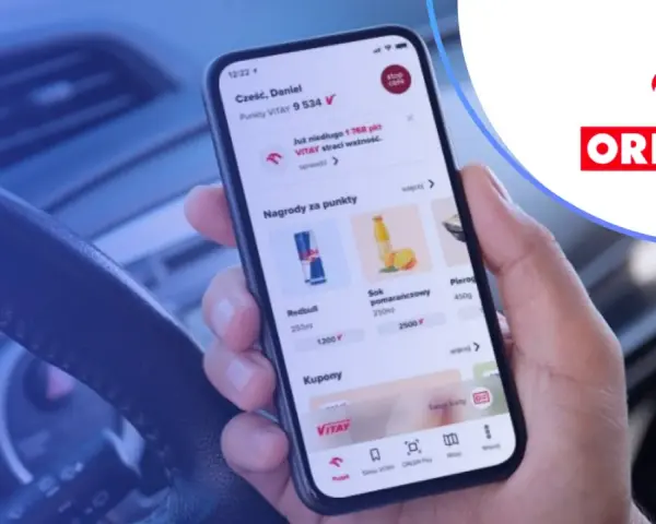 Dłoń trzyma smartfon z aplikacją ORLEN Pay, pokazującą punkty VITAY i nagrody. Kierownica samochodu w tle.