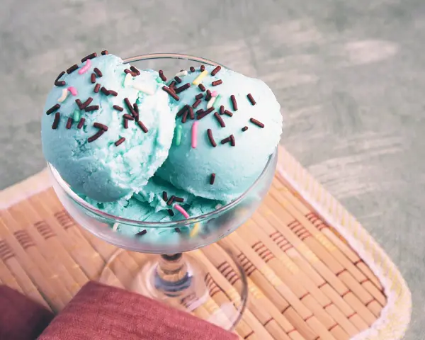 Ein Glas mit blauem Smurf-Eis, bestreut mit bunten Streuseln, steht auf einem Holzuntersetzer.