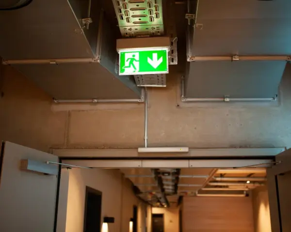 Zielone światła awaryjne wskazują drogę ewakuacji w korytarzu z widocznymi instalacjami wentylacyjnymi i betonowymi ścianami.