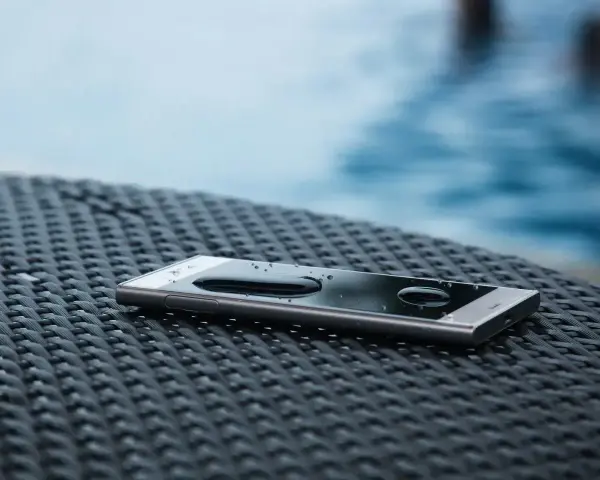 Smartfon z kroplami wody na ekranie, leżący na plecionej powierzchni. W tle widać basen. IP68 co oznacza? Ochronę przed wodą i pyłem.