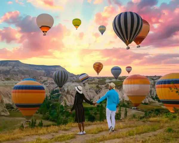 A couple holds hands, gazing at a sky filled with hot air balloons over the unique landscape of western Turkey at sunrise.