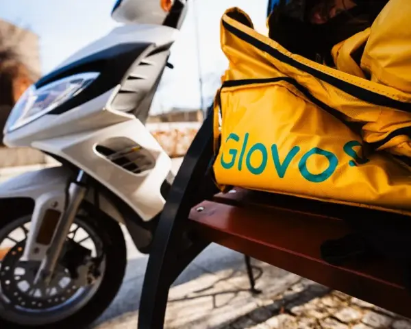 Żółta torba z logo Glovo na ławce obok białego skutera. Czeka na kolejnego kuriera Glovo.