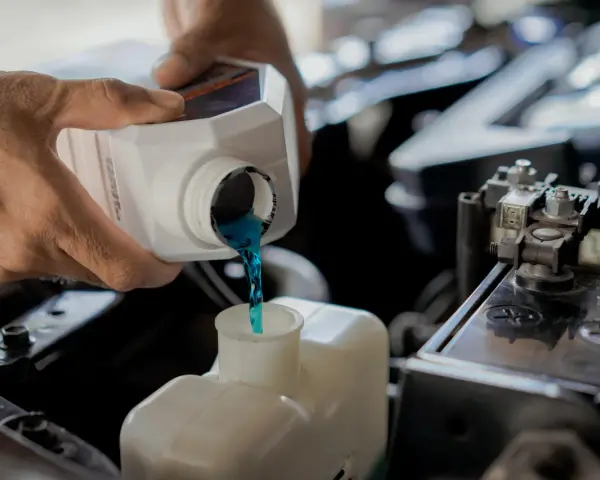 Hands pour blue coolant into a car's reservoir. This maintenance task is essential for preventing overheating, which can lead to costly car radiator replacement.