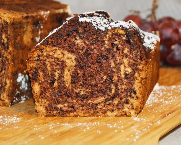 Puszysty chlebek bananowy z kakao, oprószony cukrem pudrem, prezentuje się apetycznie na drewnianej desce.
