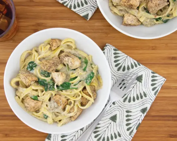 Delicioso plato de pasta alfredo con trozos de pollo y espinacas, servido en un tazón blanco sobre una tabla de madera.