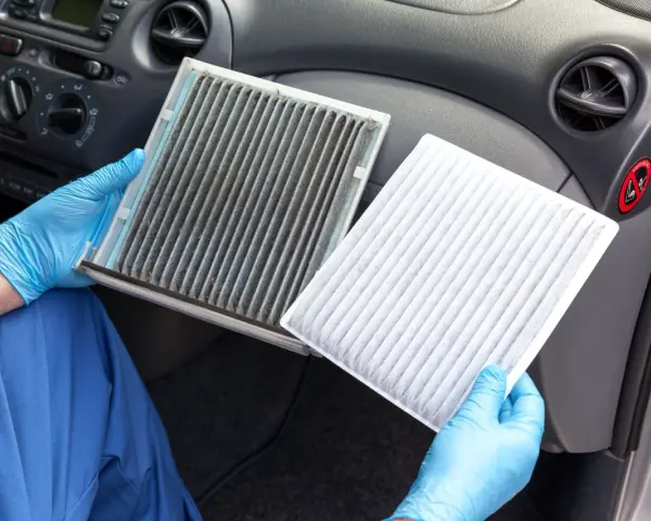 A dirty cabin air filter can make your car smell. A gloved hand holds a very dirty filter next to a clean, white one, showing the stark contrast.
