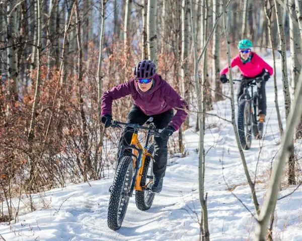 Two people enjoy a winter mtb ride on a snowy trail through a birch forest.