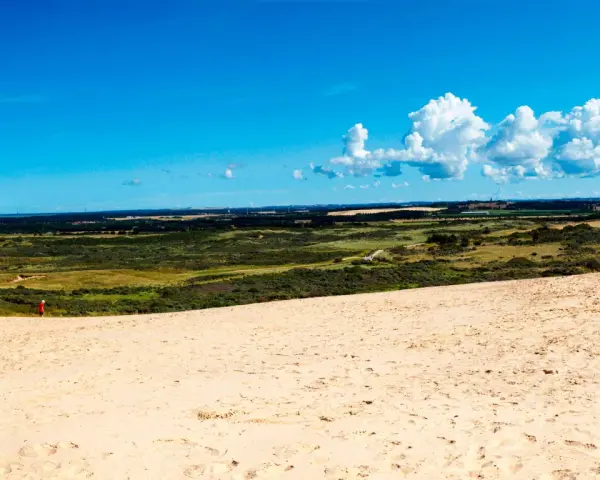 En person i röd jacka vandrar på en sanddyn vid Jyllands strand. Landskapet sträcker sig mot en blå himmel med vita moln.