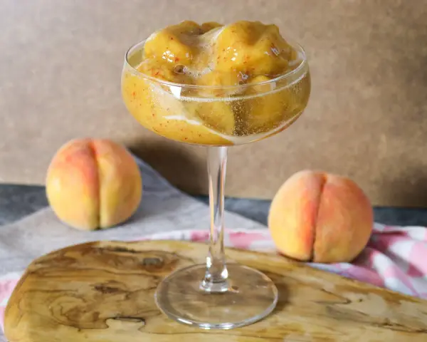 Erfrischendes Pfirsich-Prosecco-Sorbet in einem eleganten Glas, daneben reife Pfirsiche.