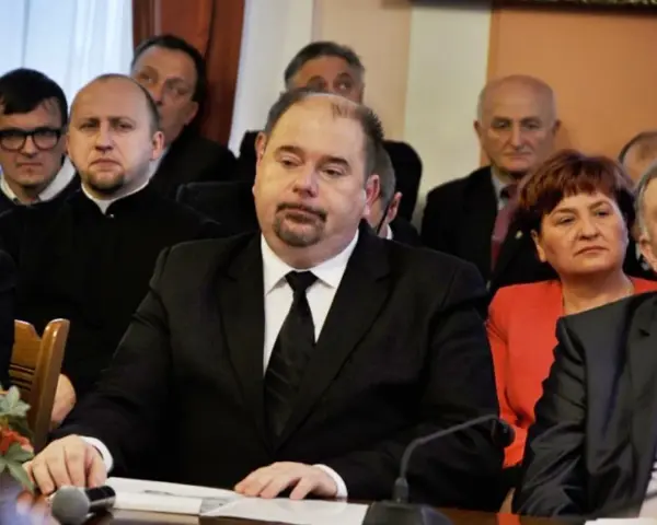 Jarosław Paluch w czarnym garniturze siedzi przy stole, otoczony innymi osobami.