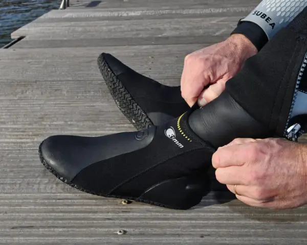 Mężczyzna zakłada czarne buty neoprenowe na drewnianym pomoście przy wodzie.