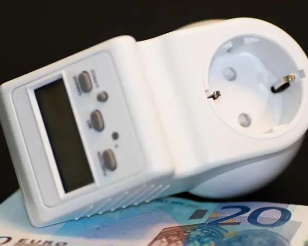 Biały miernik zużycia prądu z wyświetlaczem i przyciskami, leżący na banknocie 20 euro.