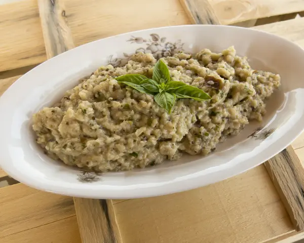 Hummus di melanzane cremoso, guarnito con basilico fresco, servito in un piatto ovale su un tavolo di legno grezzo.