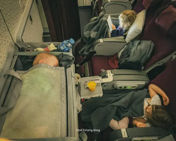 Lot z niemowlakiem i śpiącymi dziećmi w samolocie. Maluch odpoczywa w gondoli, starszaki pod kołderkami.