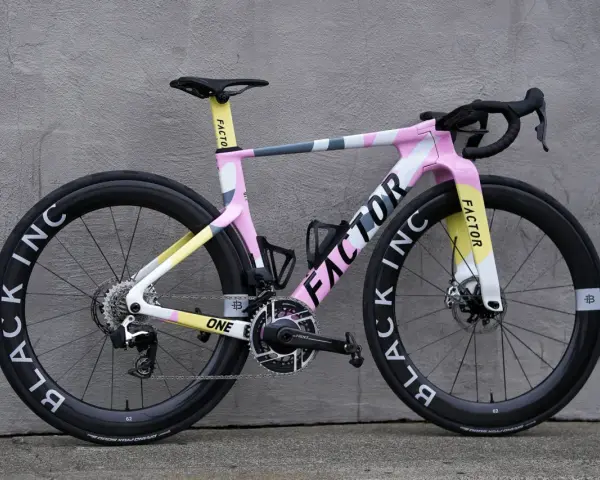 A pink, yellow, and gray Factor road bike with black wheels against a gray wall.
