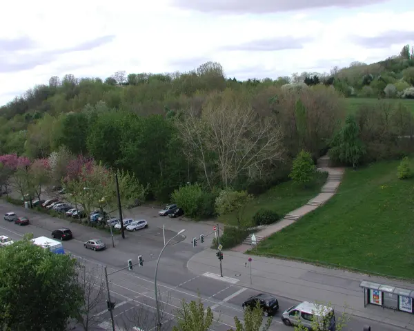 Grüne Hügel und Bäume im Volkspark Prenzlauer Berg. Autos parken am Straßenrand, eine Treppe führt einen Hang hinauf.