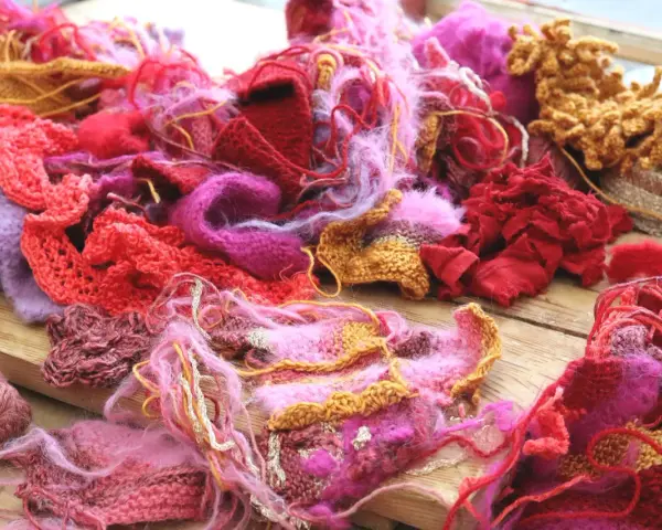 En hög av garn och stickade tygbitar i rosa, rött och lila. En del har en fin stickad kant som inte rullar sig.