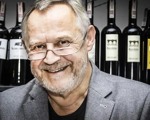 Marek Kondrat, z uśmiechem i okularami, na tle butelek wina. Jego majątek to nie tylko sukcesy zawodowe, ale i pasja do trunków.