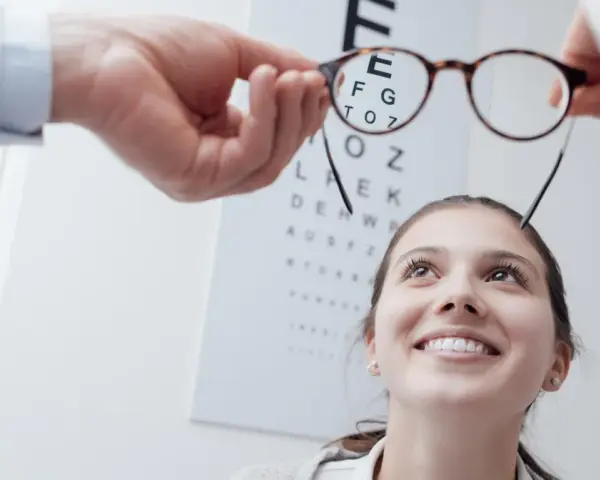 Okulista trzyma okulary przed twarzą uśmiechniętej kobiety na tle tablicy Snellena.