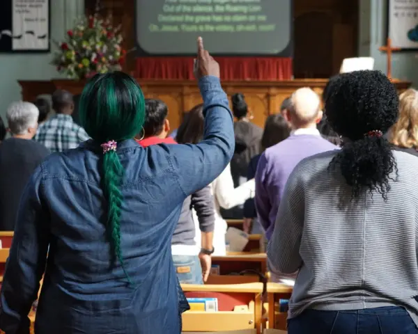 A woman with green hair raises her hand, perhaps inspired by the lyrics on screen, offering chant ideas for the congregation.
