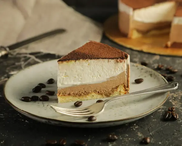 Pyszny sernik tiramisu z warstwami kremu i biszkoptu, posypany kakao. Na talerzu ziarna kawy i widelec.