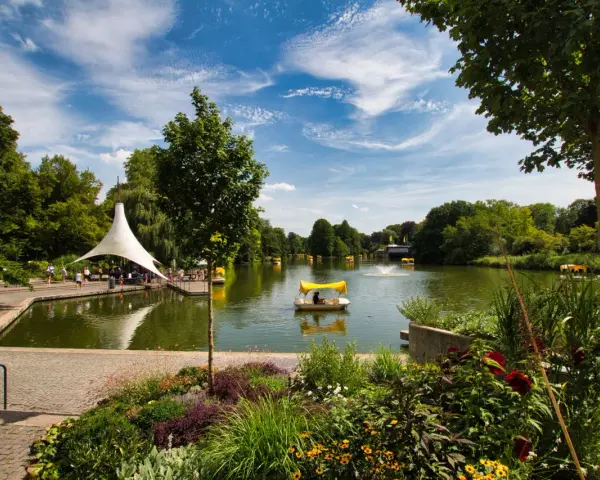 Ein sonniger Tag am Badesee Mannheim. Mehrere Tretboote mit gelben Verdecken gleiten über das Wasser, umgeben von üppigem Grün und blühenden Blumen.