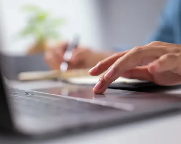 Dłoń na touchpadzie laptopa, w tle notatnik i długopis.