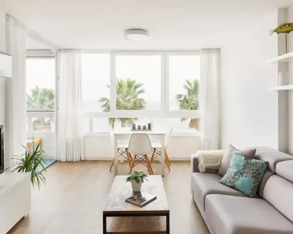 Un luminoso salón estilo nórdico con sofá gris, mesa de centro de madera y grandes ventanales con vistas a palmeras.