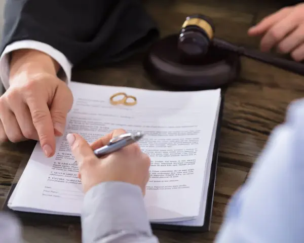 Gerichtlicher Hammer und Eheringe auf Scheidungsdokumenten. Ein Anwalt zeigt auf einen Paragraphen, während eine Person unterschreibt. Ein wichtiger Termin, der vielleicht Sonderurlaub erfordert.