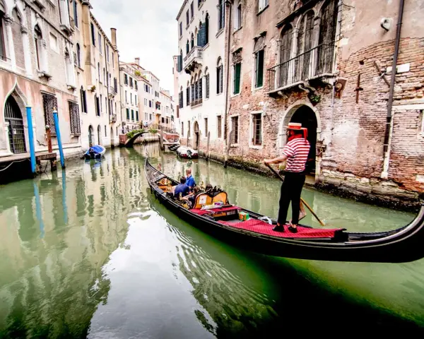 Gondoliere in abito a righe guida una gondola con turisti lungo un canale veneziano. Chissà quanto costa un giro in gondola a Venezia per vivere questa magia.
