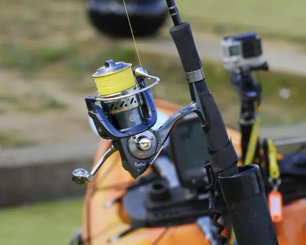 Niebieski kołowrotek spinningowy z żółtą żyłką, gotowy do akcji. W tle widać kajak i kamerę GoPro.