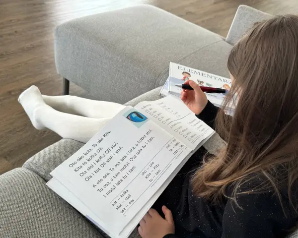 Dziewczynka z długimi włosami uczy się czytać z elementarza. Na stronach widać proste teksty, idealne do nauki czytania dla 7 latka.