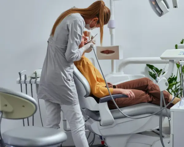 Dentystka w masce i rękawiczkach wykonuje zabieg pacjentowi siedzącemu na fotelu dentystycznym.