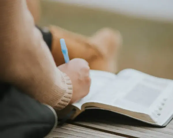 Osoba pisze w otwartej księdze, być może dziennik. W tle widać fragmenty ubrania. To moment refleksji, gdzie żal doskonały może znaleźć swoje ujście w słowach.