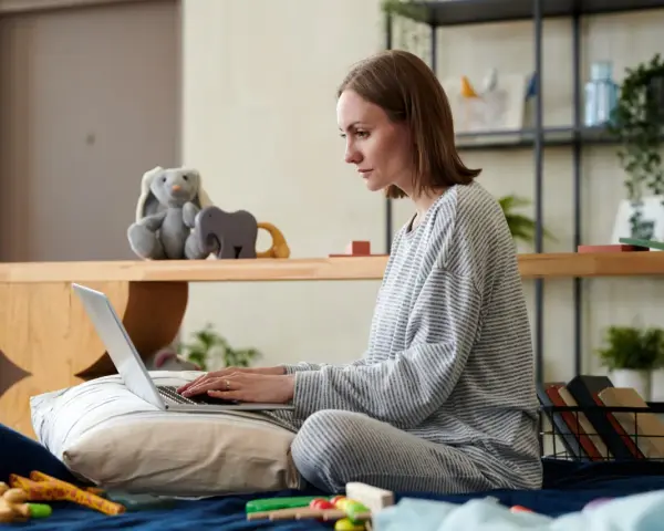 Kobieta w piżamie pisze na laptopie, przygotowując wniosek o urlop macierzyński. Obok pluszowy królik i zabawki.
