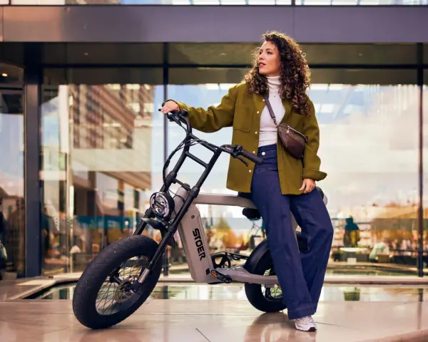 En kvinna med lockigt hår sitter på en STOER elcykel framför en modern byggnad. Hon bär en grön jacka och jeans.