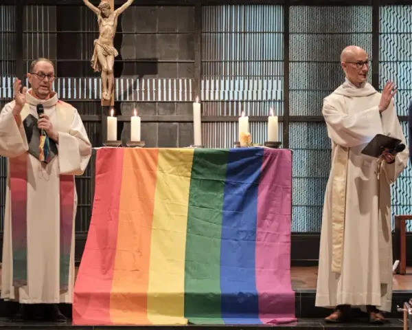 Zwei Geistliche stehen vor einem Altar mit einer Regenbogenfahne. Die Kirche feiert die gleichgeschlechtliche Ehe.