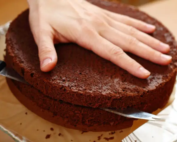 Eine Hand hält einen Schokoladenkuchen, während ein Messer ihn waagerecht durchschneidet, um den tortenboden zu teilen.