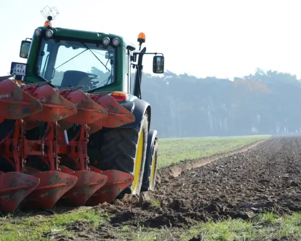 Zielony traktor z pługiem orze pole. W tle mglisty las.
