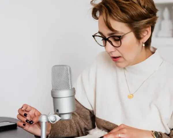 Mujer con gafas lee un libro frente a un micrófono, aprendiendo como escribir mejor.