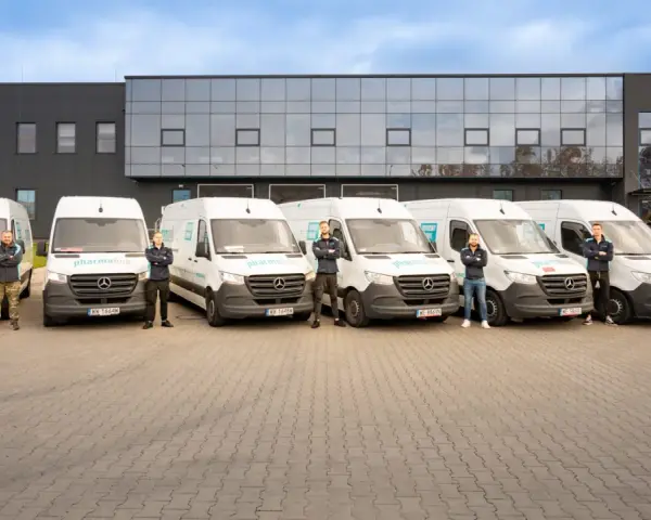 Sześciu mężczyzn w uniformach stoi przed rzędem białych vanów z logo "pharmalink". To zespół odpowiedzialny za transport leków, profesjonalny przewoźnik.