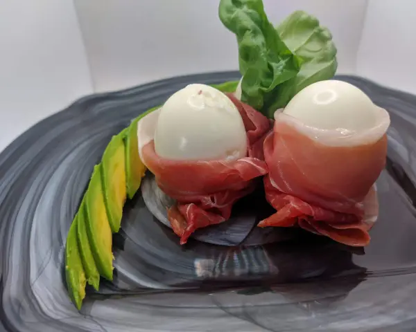 Due uova di quaglia sode avvolte in prosciutto crudo, servite con fette di avocado e foglie verdi su un piatto scuro.