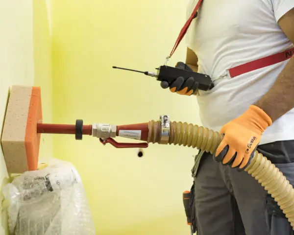Un técnico usa un dispositivo para insuflar aire a través de una manguera y una esponja en una pared amarilla.