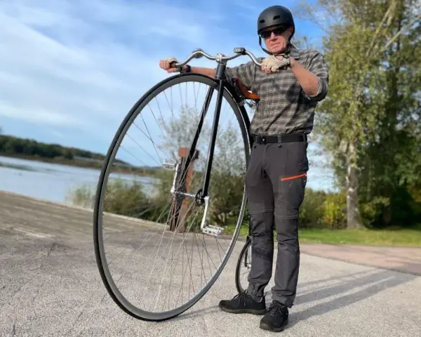 Man med hjälm och solglasögon står bredvid en cykel med stort framhjul vid en sjö.