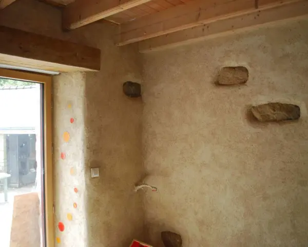 Un mur avec un enduit chaux finition texturé, intégrant des pierres apparentes et des touches de couleur. Le bois des poutres et du cadre de fenêtre apporte de la chaleur.