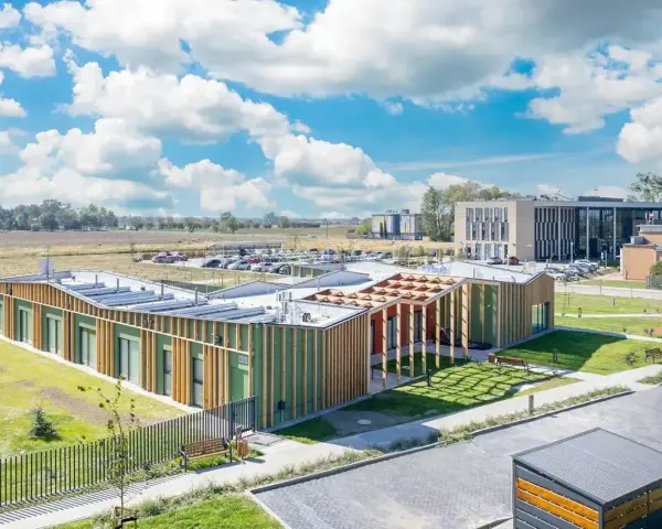 Nowoczesny budynek użyteczności publicznej z zielonymi fasadami i drewnianymi elementami, otoczony trawnikami i parkingiem.